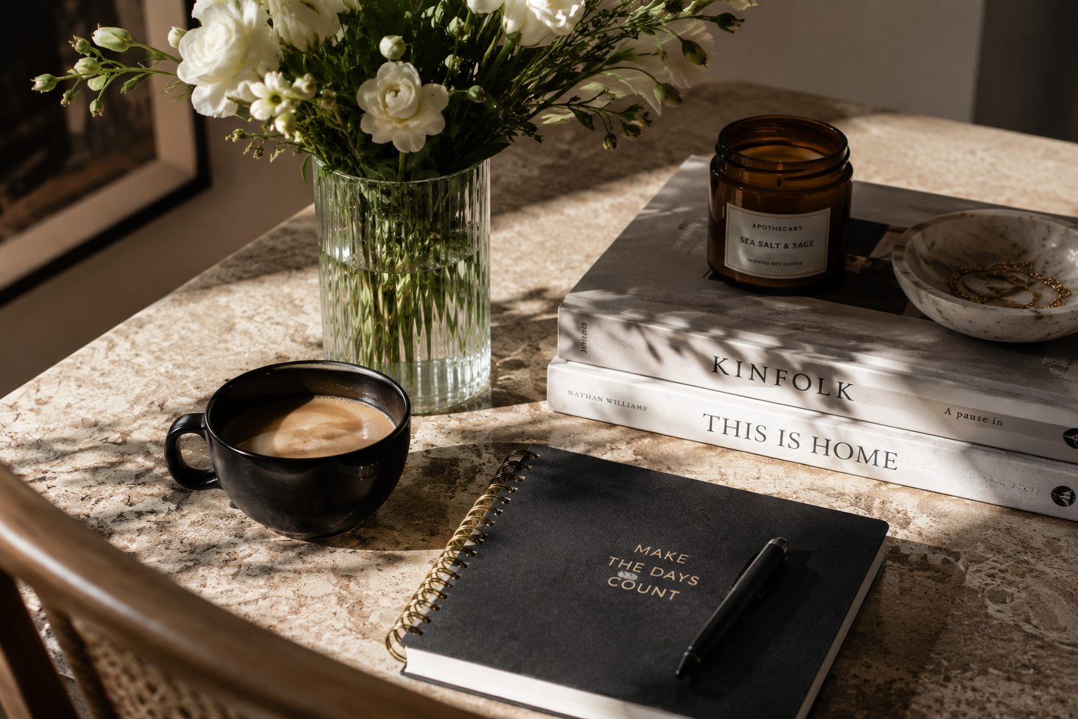 Editorial-style still life featuring a cup of coffee, fresh white flowers, a journal, and books on a sunlit table, representing simple personal traditions that make ordinary weeks more meaningful