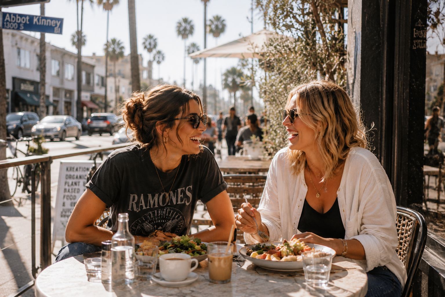 Two women wearing sunglasses laugh and enjoy lunch together at an outdoor café on a sunny California city street lined with palm trees.
